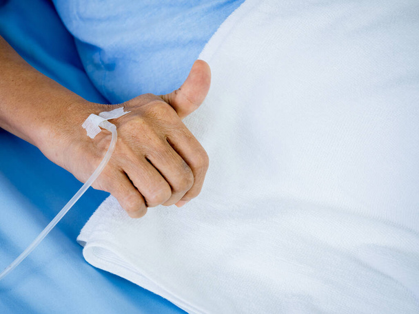 Закройте солевой раствор в руках пожилых пациентов с большим пальцем в синем платье, лежащем на кровати с белым одеялом в больничной палате с копировальным местом. Рука пожилого пациента получает физраствор внутривенно. - Фото, изображение