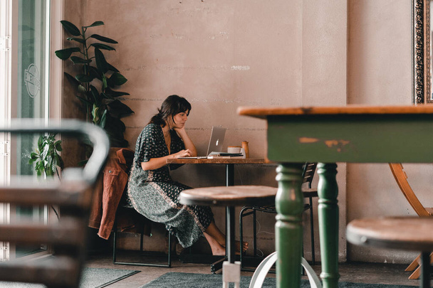 young caucasian woman with disheveled hair in long dress sitting on metal chair by wall typing laptop at wooden table in quiet coffee shop, the mylk bar, new zealand - Lifestyle concept - Photo, Image
