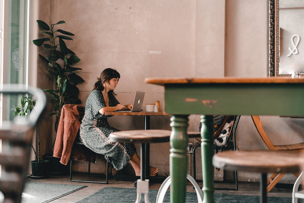 serious concentrated caucasian girl in long gray dress sitting working with laptop at wooden table indoors in calm and nice coffee shop, the mylk bar, new zealand - Lifestyle concept - Foto, Bild