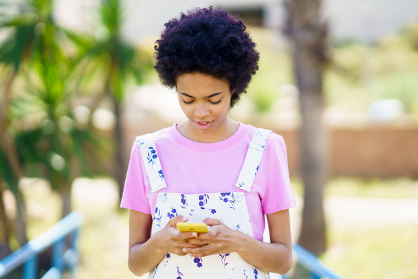Feliz mujer afroamericana en ropa casual desplazándose por las redes sociales en el teléfono celular mientras está de pie en la calle con árboles contra el fondo borroso - Foto, imagen