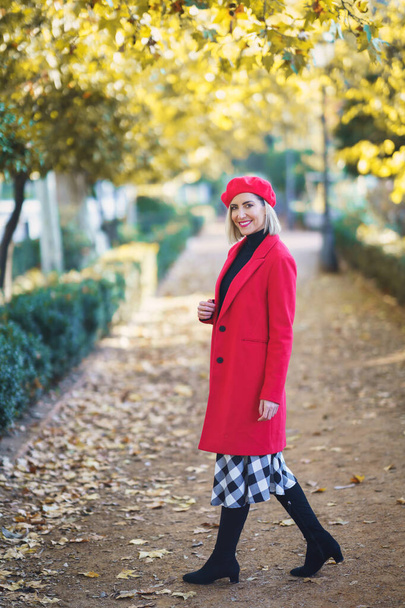 A vidám, vörös ruhás nő egész teste a kamerába néz, miközben ősszel a parkban áll. - Fotó, kép