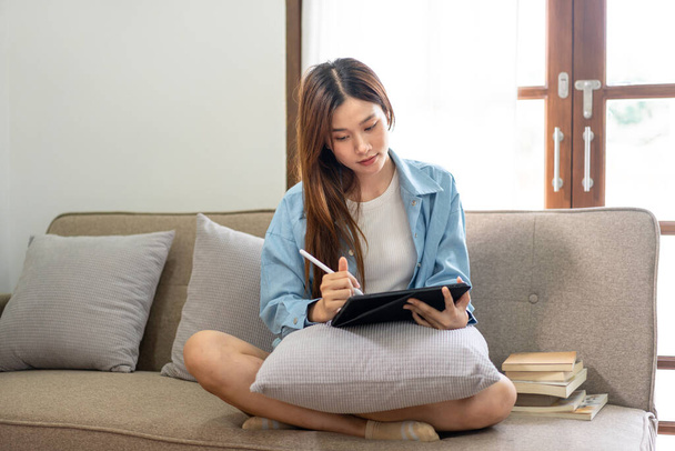 Молода жінка аналізує стратегічну мету бізнесу і робить нотатки на планшеті, працюючи над зручним диваном у вітальні вдома
. - Фото, зображення