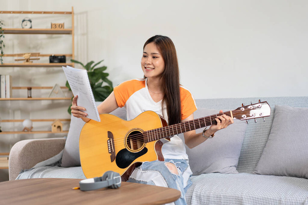 Молода жінка тримає книгу пісень і акордів, щоб грати на гітарі і тренуватися співати пісню, сидячи на зручному дивані у вітальні вдома
. - Фото, зображення