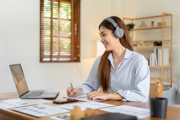 Female freelance is reading business data on laptop and writing information on notebook while wearing headphone to listening music and working on the table in living room at home office. - Photo, Image