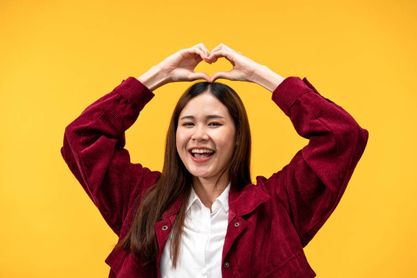 Jovem mulher asiática vestindo jaqueta vermelha e grande sorriso ao colocar uma forma de coração com as mãos na cabeça isolada sobre fundo amarelo. - Foto, Imagem