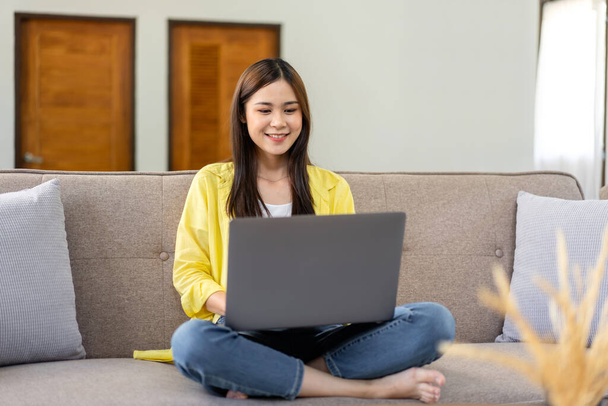 Egy fiatal nő guggolva ül a kényelmes kanapén, és laptopot tesz a lábára, hogy üzleti jelentést írjon, miközben otthon dolgozik a nappaliban.. - Fotó, kép