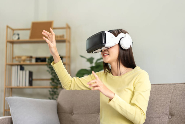 Молодая азиатка в очках виртуальной реальности смотрит видео и играет в игры с виртуальным опытом, используя руки с пальцами, чтобы прикоснуться к воздуху и сидеть на диване в гостиной. - Фото, изображение