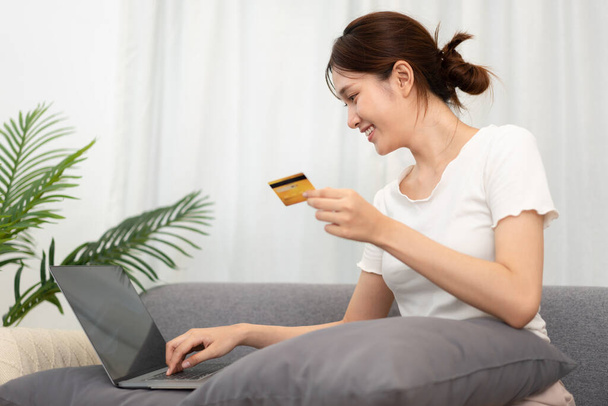 Teenage girl is holding credit card and typing credit card number for payment after shopping online on laptop while sitting to relax comfortable on the couch in living room. - Fotó, kép