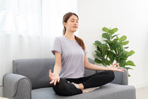 Young woman is relaxing into the lotus position for meditation on couch before workout yoga exercise in her living room at home. - Zdjęcie, obraz