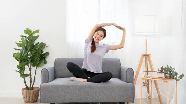 Νεαρή γυναίκα κάθεται να τεντώσει τα χέρια της για χαλάρωση στον καναπέ μετά την άσκηση γιόγκα προπόνηση στο σαλόνι της στο σπίτι. - Φωτογραφία, εικόνα