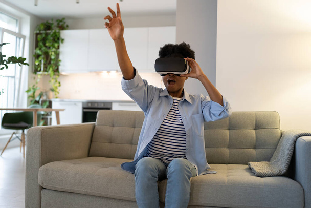 Zachęcona Afroamerykanka korzystająca z okularów wirtualnej rzeczywistości siedzi na szarej sofie we własnym mieszkaniu. Czarna dziewczyna w swobodnym ubraniu korzysta z technologii VR, aby oglądać filmy 3D lub zanurzyć się w metawersie - Zdjęcie, obraz