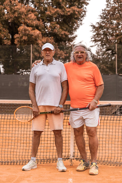 portrait of two senior tennis players dressed in sportswear posing at the end of the game on a clay tennis court - retired wellness concept - Fotografie, Obrázek