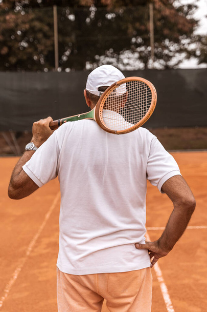 a retired tennis player dressed in sportswear posing with his back to the camera on a clay tennis court - retired wellness concept - Foto, Bild