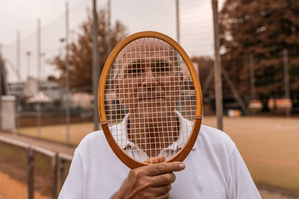 portrait of senior tennis player dressed in sportswear posing with racket on face on a clay tennis court - retired wellness concept - Fotografie, Obrázek