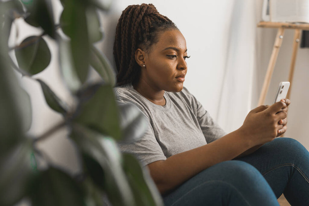 Πορτρέτο μιας εμφανίσιμης αφρικανίδας φοιτήτριας ντυμένης άνετα με κινητό τηλέφωνο και δακτυλογραφώντας μηνύματα και επικοινωνώντας με φίλους στα κοινωνικά δίκτυα χρησιμοποιώντας υψηλή σύνδεση στο Διαδίκτυο. - Φωτογραφία, εικόνα