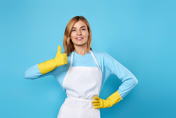 smiling blonde woman in yellow rubber gloves showing ok gesture on blue background. cleaning - Fotografie, Obrázek