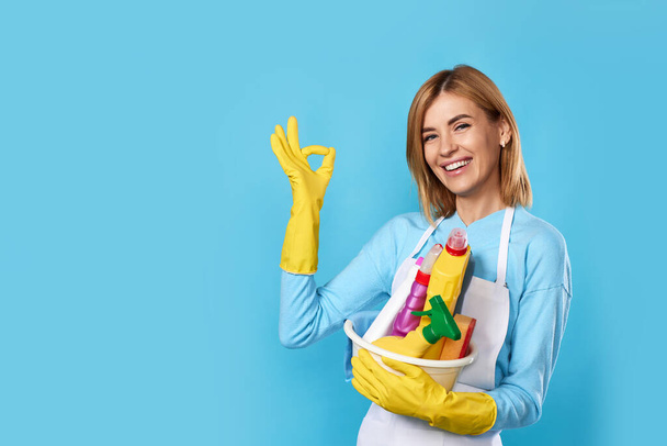 happy blonde woman in gloves and cleaner apron holding bucket of detergents and showing Ok gesture on blue background. - Фото, изображение