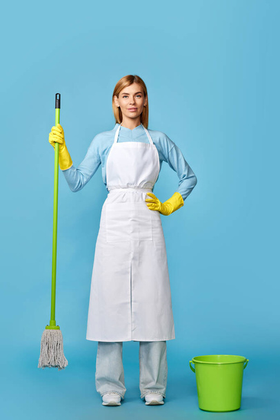 professional cleaner blonde woman in rubber gloves and cleaner apron washing floor with mop on blue background. Full length - Fotoğraf, Görsel