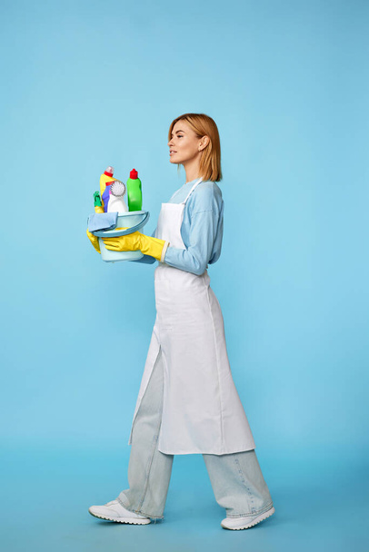 happy cheerful blonde woman in rubber gloves and cleaner apron with cleaning supplies on blue background. Full length - Φωτογραφία, εικόνα