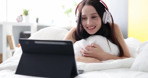 Sonriendo mujer feliz en los auriculares se comunica por videollamada mientras está acostado en la cama. Concepto positivo de comunicación en línea - Metraje, vídeo