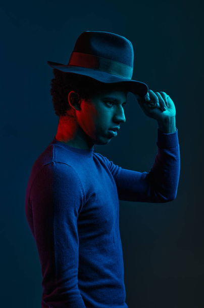 Young man in hat with stylish outlook over dark studio background under neon light illumination - Фото, изображение