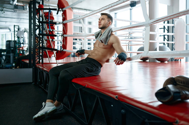 Portret van gespierde mannelijke bokser zittend op boksring mat en rust na harde training of competitieve wedstrijd - Foto, afbeelding