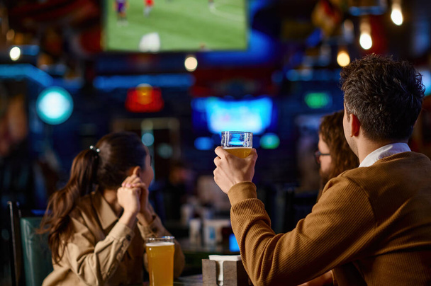 Arkadaş grubunun yakın çekimleri, bira barında birlikte otururken bira ile kadeh kaldırması. Bardağı soğuk içecekle tutan adama odaklan. - Fotoğraf, Görsel