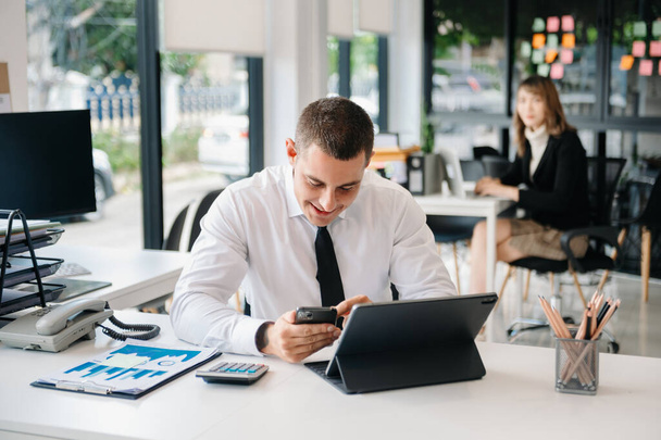 Young business man working at modern office with smartphone, tablet and taking notes on the paper. woman on background  - Zdjęcie, obraz