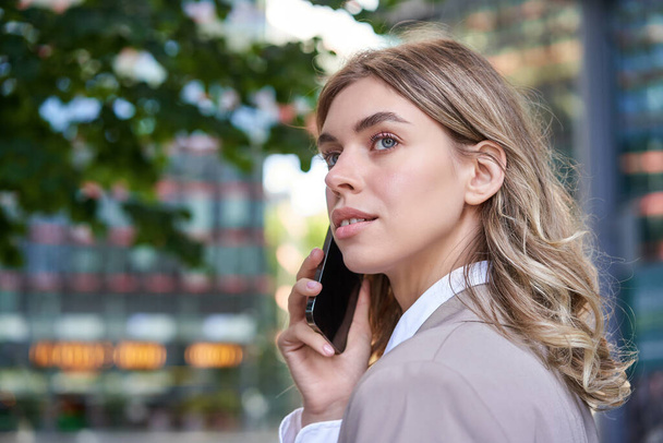 Close up shot of businesswoman talking on mobile phone. Corporate woman calling someone, looking around, standing outdoors. - Photo, Image