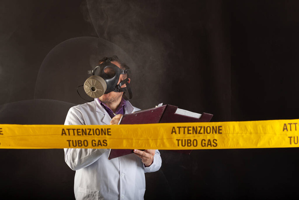 Lékařský inženýr drží složku s dokumenty, dělá si poznámky, nosí chemické ochranné oděvy a Antigas Mask řídí nouzové situace v chaosu během úniku plynu. Na žluté pásce je napsáno upozornění "Trubka s plynem" - Fotografie, Obrázek