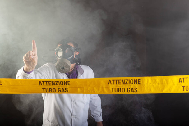 Ιατρικός μηχανικός που φορούσε μάσκα αντιαερίων κατά τη διάρκεια της κρίσης διαρροών αερίου και της έκτακτης ανάγκης κατά τη διάρκεια του χάους σε ένα μαύρο δωμάτιο με καπνό. Στην κίτρινη ταινία η γραπτή ειδοποίηση "προσοχή σωλήνα αερίου" - Φωτογραφία, εικόνα