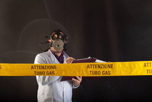 Un ingeniero médico sostiene la carpeta de documentos tomando notas, vistiendo ropa de protección química y Antigas Mask dirige la emergencia en el caos durante las fugas de gas. En la cinta amarilla el aviso escrito "tubo de gas de atención" - Foto, imagen
