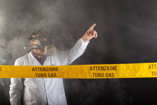Ένας ιατρικός μηχανικός με προστατευτική χημική ενδυμασία και μάσκα antigas κατευθύνει την κατάσταση έκτακτης ανάγκης στο χάος κατά τη διάρκεια των διαρροών αερίου. Στην κίτρινη ταινία η γραπτή ειδοποίηση "προσοχή σωλήνα αερίου" - Φωτογραφία, εικόνα