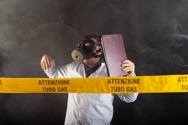 Ένας ιατρικός μηχανικός κρατά το φάκελο εγγράφων κρατώντας σημειώσεις, φορώντας χημική προστατευτική ενδυμασία και μάσκα antigas κατευθύνει την κατάσταση έκτακτης ανάγκης στο χάος κατά τη διάρκεια των διαρροών αερίου. Στην κίτρινη ταινία η γραπτή ειδοποίηση "προσοχή σωλήνα αερίου" - Φωτογραφία, εικόνα