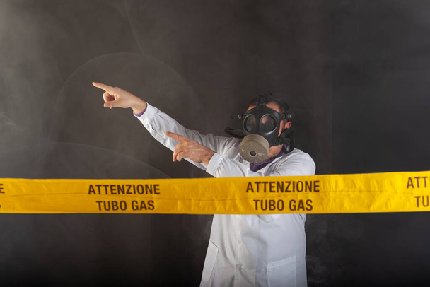 Ένας ιατρικός μηχανικός με προστατευτική χημική ενδυμασία και μάσκα antigas κατευθύνει την κατάσταση έκτακτης ανάγκης στο χάος κατά τη διάρκεια των διαρροών αερίου. Στην κίτρινη ταινία η γραπτή ειδοποίηση "προσοχή σωλήνα αερίου" - Φωτογραφία, εικόνα