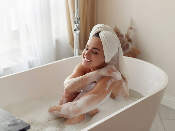 Закінчила щасливу кавказьку жінку, що сиділа в ванній з піною, миючи своє тіло і посміхаючись у доброму настрої. Ранковий режим гігієни. Прекрасна молода жінка приймає спокійну ванну. Самодогляд - Фото, зображення