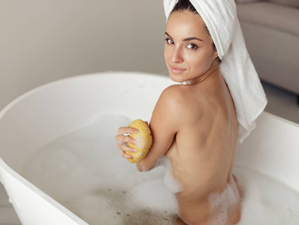 Visão superior jovem mulher atraente contente sentado nu na banheira em água com espuma de sabão braço de banho esfregando com esponja. Fecha. Caucasiano corpo de lavagem feminino feliz olhando para a câmera. - Foto, Imagem