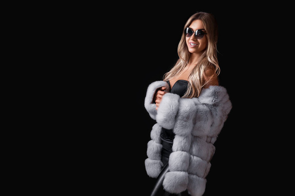 Beautiful atrractive blonde girl in sunglasses posing on a black background in a fur coat. Commercial concept. Studio shot.  - Fotó, kép