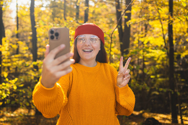 Портрет счастливой улыбающейся молодой женщины в красной шляпе, разговаривающей с помощью мобильного телефона, делающей селфи, гуляющей в одиночестве в осеннем парке на природе осенью. Селективный фокус.  - Фото, изображение