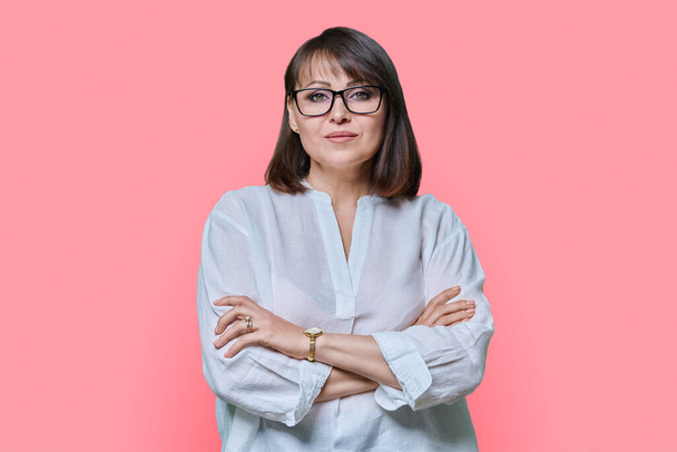 Serious confident middle aged woman in glasses on pink color background. Experienced mature female in white casual clothes looking at camera. Lifestyle, confidence, calmness, mature people concept - 写真・画像