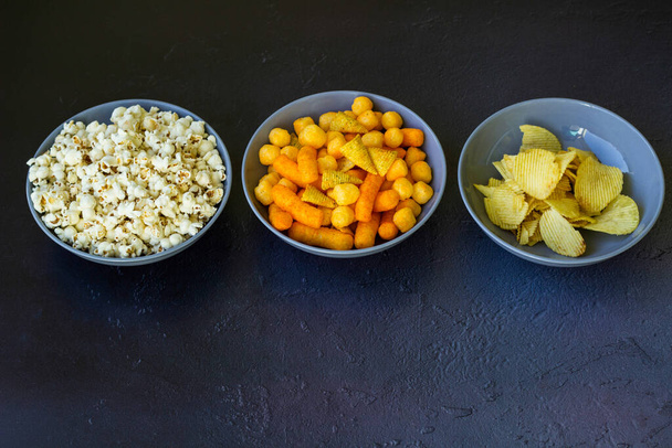 Salty snacks assortment shot on dark slate table. The picture includes potato chips, popcorn, peanut, cheese sticks, ketchup sticks, tzatziki chips, smoky flips, and others - Fotoğraf, Görsel
