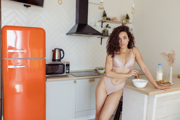 Attractive young beautiful brunette woman at home standing in kitchen near counter during breakfast looking at camera. Pretty female in sexy underwear eating cereals indoors. Domestic routine. - Photo, Image