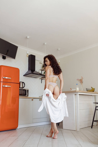 Back view of pretty hot housewife in lingerie at home standing in the kitchen taking off white shirt. Rear view. Low angle view. Caucasian beautiful female in sexy underwear indoors. Domestic life - Photo, image
