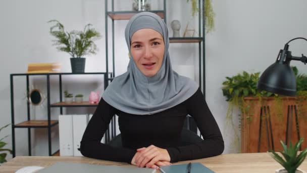 Muzułmańska bizneswoman w hidżabie pracująca na laptopie uśmiechnięta przyjazna przed kamerą i gestykulująca cześć, cześć, assalamu alaikum, powitanie, powitanie z gościnnym wyrazem twarzy przy biurku - Materiał filmowy, wideo
