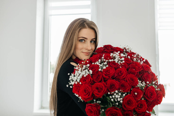 Όμορφη κοπέλα με μακριά μαλλιά με ένα τεράστιο μπουκέτο τριαντάφυλλα χαμογελά και κοιτάζει στην κάμερα. Λουλούδια ως δώρο και παράδοση των λουλουδιών για το κάθε διακοπές. Ημέρα του Αγίου Βαλεντίνου - Φωτογραφία, εικόνα