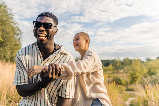 Sranda a smích s přáteli a blízkými. Multietnický pár se baví na venkově. Smějící se černoch v neformálním oblečení, dotýkající se paže svého plešatého bělocha. Kvalitní fotografie - Fotografie, Obrázek
