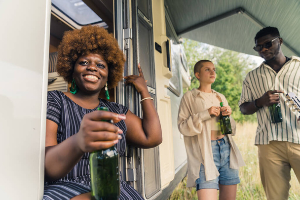 Owner of summer traveling van - beautiful smiling Black woman sitting at the door with one green glass beer bottle in hand. Multiracial friends in the background. Afternoon leisure relax. High quality - Photo, Image