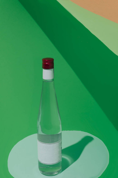 Botella sobre fondo verde y sombras. Obturador rojo. Etiqueta en blanco. Entorno conceptual, minimalismo. - Foto, Imagen