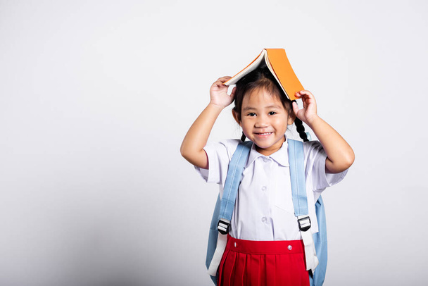 Asian adorable toddler smiling happy wearing student thai uniform red skirt stand holding book over head like roof in studio shot isolated on white background, Portrait little children girl preschool - Zdjęcie, obraz