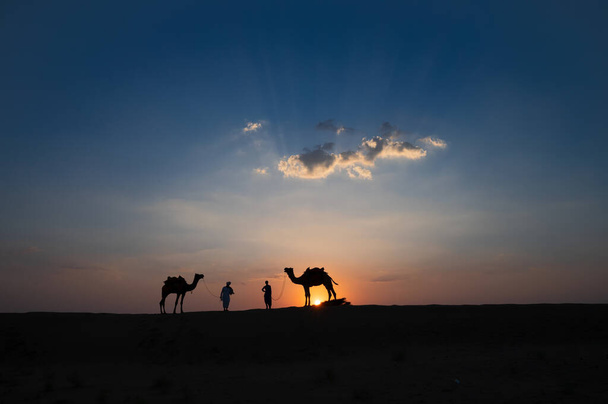 インドのラジャスタン州サール砂漠の砂丘で2人のカメラマンとラクダのシルエット。背景に夕日と雲、空。ラクダ乗りで観光客で生計を立てる. - 写真・画像